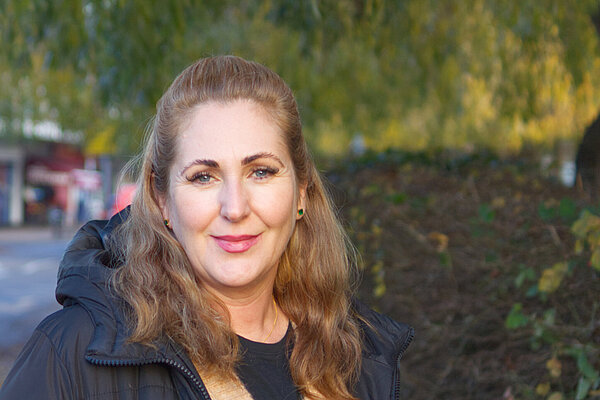 Mia Handley faces the camera. The green hedges and willow tree of Little Reddings School make up most of the background. The shops are off in the distance.