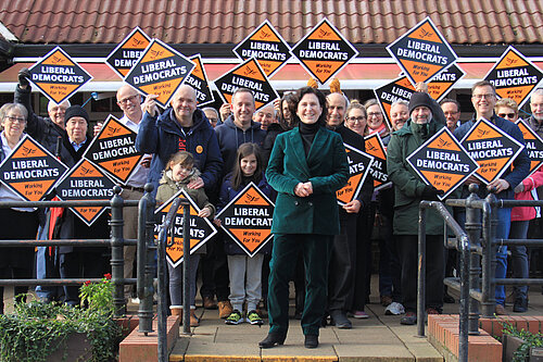 Emma Matanle in front of a large number of Hertsmere Lib Dems and their families, many are holding orange Lib Dem diamond posterboards