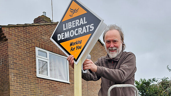 A volunteer on a ladder is putting up a Lib Dem garden posterboard 