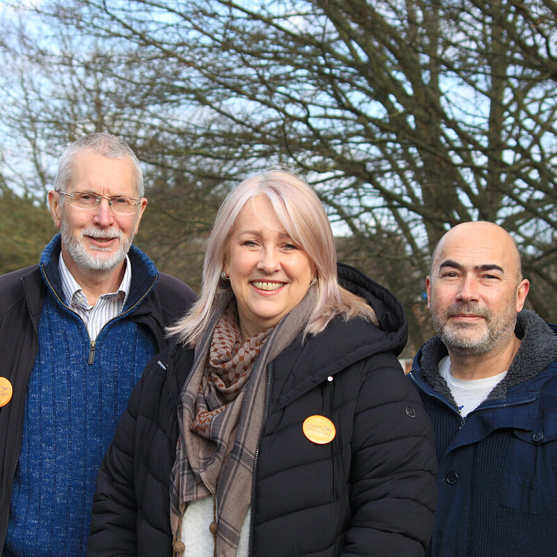 Alan Mathews, Louise Nicolas and Paul Richards stand together on a winter's day with bare trees behind them. Two of them are wearing Lib Dem badges