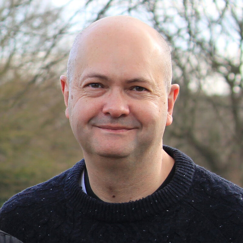 Photo of Maxie Allen, Vice Chair of the Hertsmere Lib Dems, smiling in front of some trees.