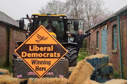 Ed Davey smashes the blue wall Ed Davey waves from tractor with Lib Dem sign, having smashed blue wall of bales